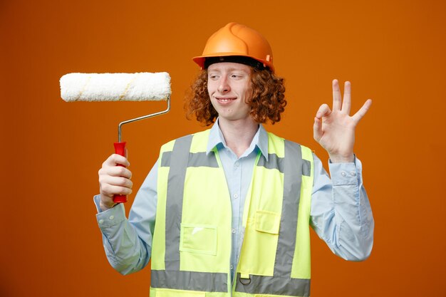 Builder young man in construction uniform and safety helmet holding paint roller looking at it with smile on face showing ok sign standing over orange background