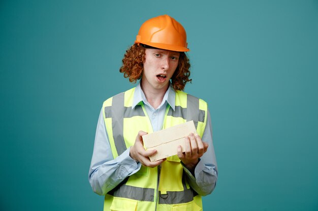 Builder young man in construction uniform and safety helmet holding two bricks looking at camera confused standing over blue background