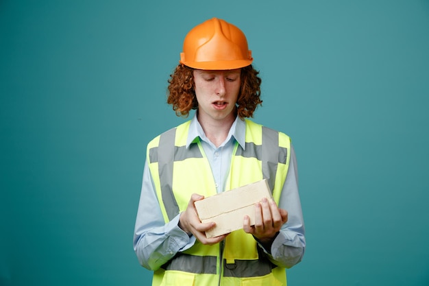 Молодой человек-строитель в строительной форме и защитном шлеме, держащий два кирпича, смотрит на него в замешательстве, стоя на синем фоне