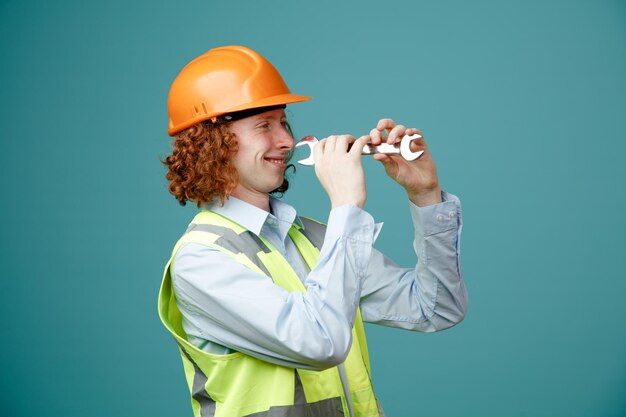 Giovane costruttore in uniforme da costruzione e casco di sicurezza con chiave inglese come piffero divertendosi sorridendo allegramente in piedi su sfondo blu