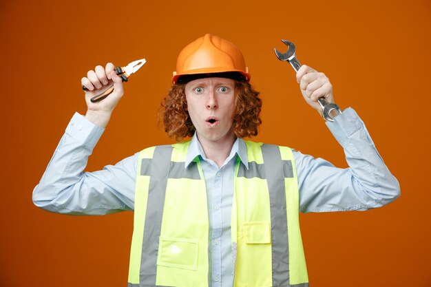 Builder young man in construction uniform and safety helmet holding wrench and pliers looking at camera amazed and surprised standing over orange background