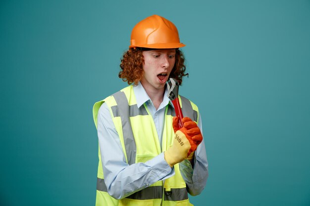 Builder young man in construction uniform and safety helmet holding wrench using as microphone having fun singing standing over blue background