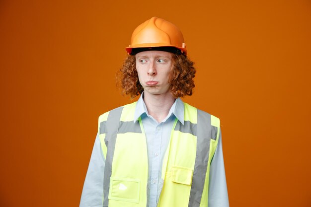 Builder young man in construction uniform and safety helmet looking aside being displeased with sad expression making wry mouth standing over orange background