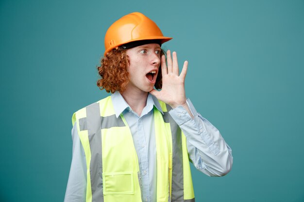 Builder young man in construction uniform and safety helmet looking aside calling someone with hand near his mouth standing over blue background