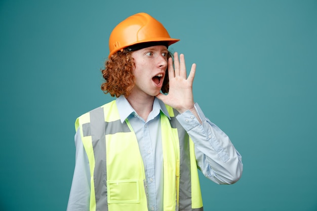 Молодой человек-строитель в строительной форме и защитном шлеме смотрит в сторону и зовет кого-то рукой у рта, стоящей на синем фоне