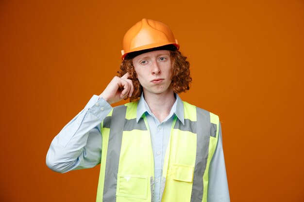 Builder young man in construction uniform and safety helmet looking aside puzzled standing over orange background