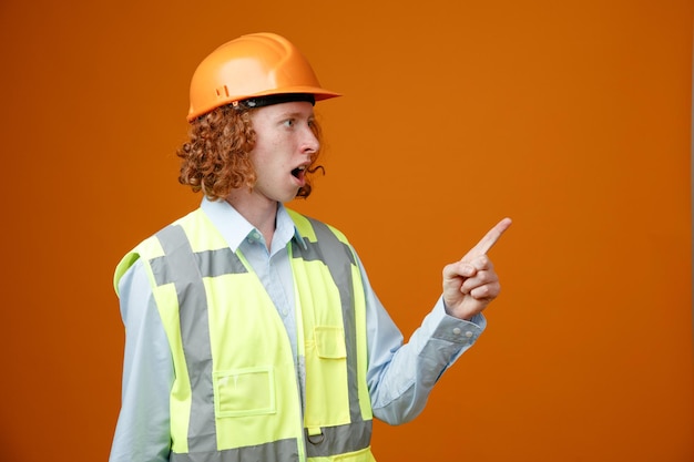 Builder young man in construction uniform and safety helmet looking aside surprised pointing with index finger at something standing over orange background