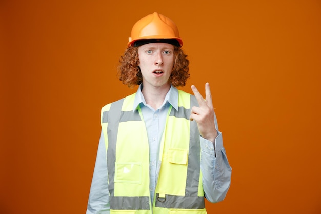 Builder young man in construction uniform and safety helmet looking at camera confused showing number two with fingers standing over orange background