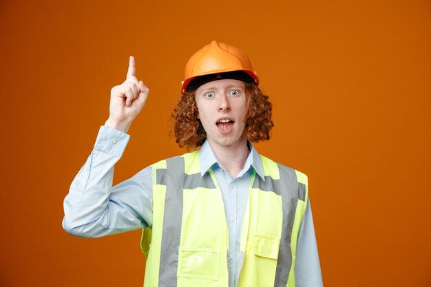 Молодой человек-строитель в строительной форме и защитном шлеме смотрит в камеру счастливым и взволнованным, показывая указательный палец, имея отличную идею, стоя на оранжевом фоне