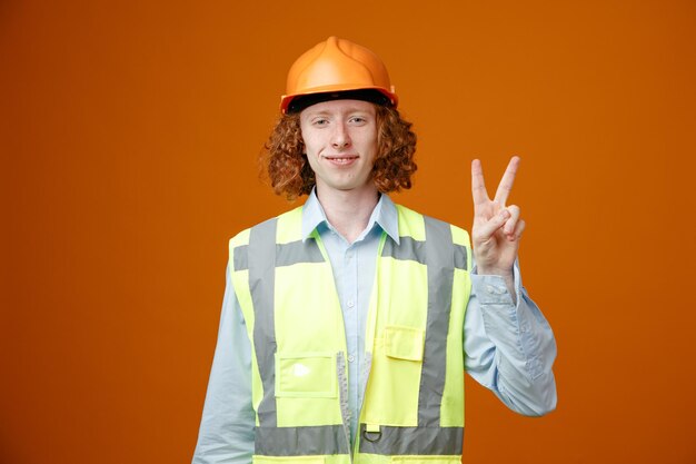 Builder young man in construction uniform and safety helmet looking at camera happy and positive smiling cheerfully showing vsign standing over orange background