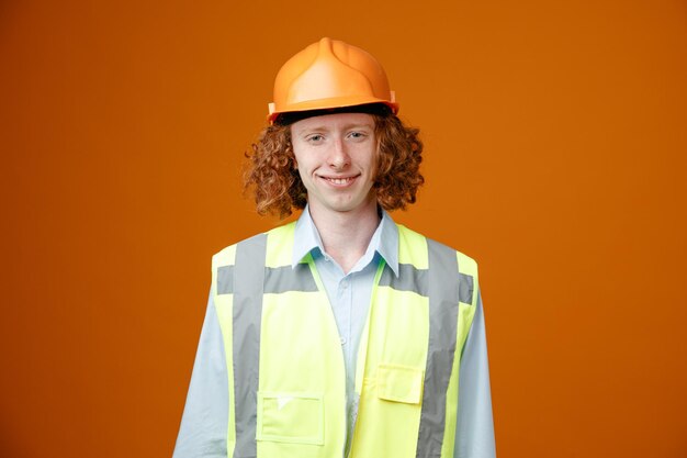 건설 유니폼을 입고 안전 헬멧을 쓴 빌더 청년은 주황색 배경 위에 즐겁게 서서 카메라를 바라보며 행복하고 긍정적인 미소를 짓고 있습니다.
