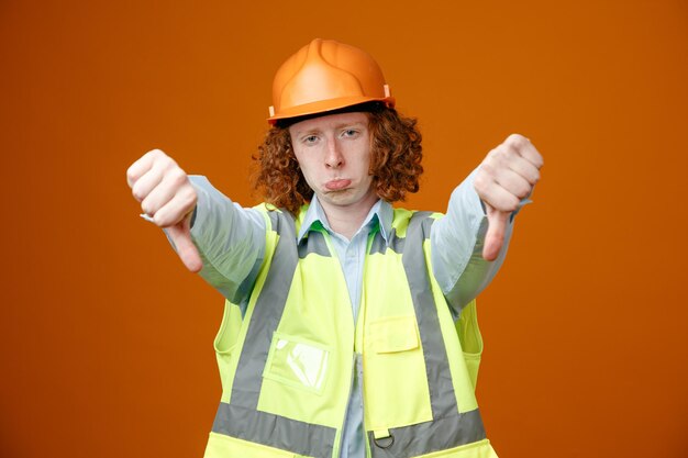 Молодой человек-строитель в строительной форме и защитном шлеме смотрит в камеру, кривя рот, показывая большие пальцы вниз, недовольный стоя на оранжевом фоне