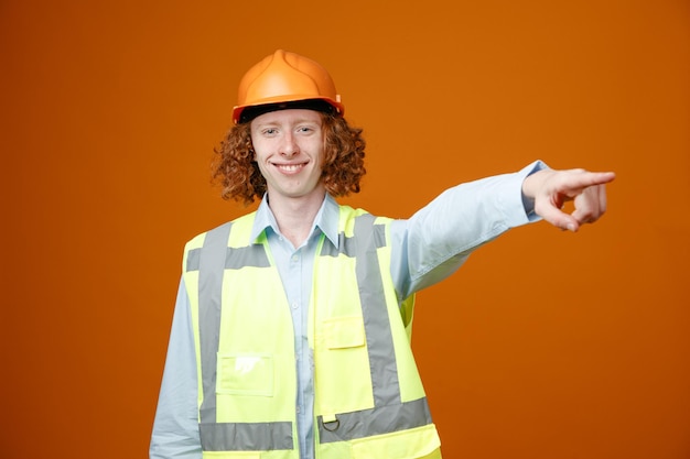 Foto gratuita giovane costruttore in uniforme da costruzione e casco di sicurezza guardando la fotocamera sorridente allegramente puntando con il dito indice a qualcosa in piedi su sfondo arancione