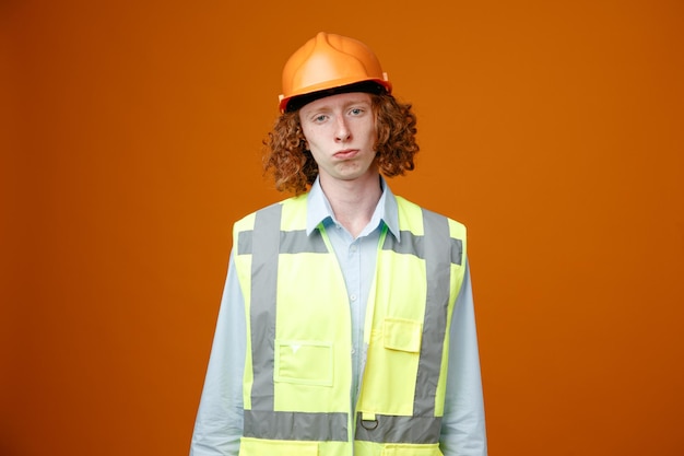 Builder young man in construction uniform and safety helmet looking at camera with sad expression making wry mouth standing over orange background