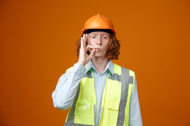 건설 유니폼을 입고 안전 헬멧을 쓴 빌더 청년은 주황색 배경 위에 지퍼가 서 있는 입을 닫는 것처럼 진지한 얼굴로 카메라를 바라보고 있습니다.