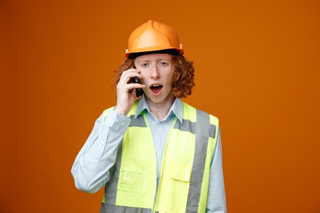 Builder young man in construction uniform and safety helmet looking surprised while talking on mobile phone standing over orange background