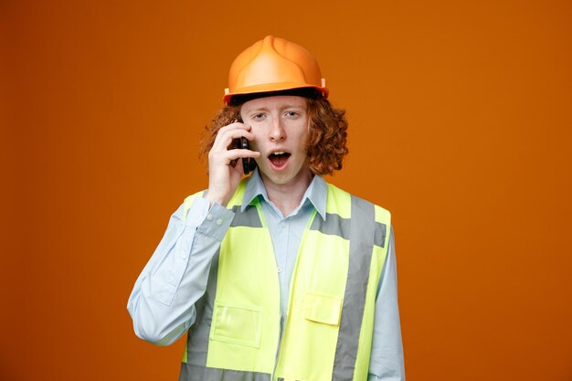 Молодой человек-строитель в строительной форме и защитном шлеме выглядит удивленным, разговаривая по мобильному телефону, стоя на оранжевом фоне