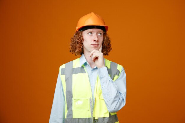 Builder young man in construction uniform and safety helmet looking up with pensive expression thinking standing over orange background