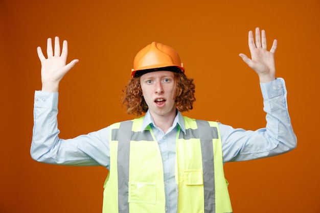 Builder young man in construction uniform and safety helmet raising arms in surrender looking confused standing over orange background