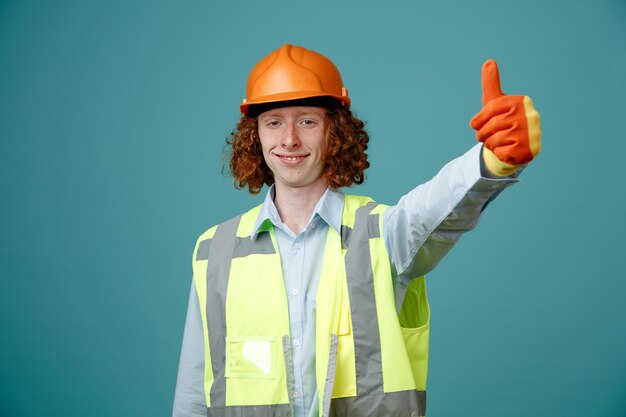 Молодой человек-строитель в строительной форме и защитном шлеме в резиновых перчатках показывает большой палец вверх счастливый и позитивно улыбающийся, стоя на синем фоне