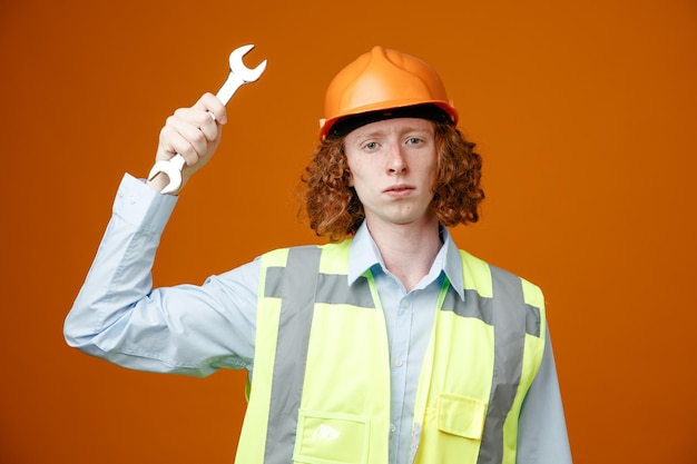 オレンジ色の背景の上に立っている深刻な顔でカメラを見てレンチを保持している建設制服と安全ヘルメットのビルダーの若い男 Premium写真