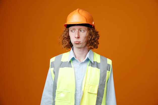 Бесплатное фото Молодой человек-строитель в строительной форме и защитном шлеме смотрит в сторону, недовольный грустным выражением лица и кривым ртом, стоящим на оранжевом фоне
