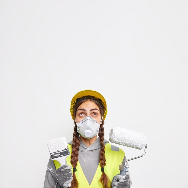 建物と建設のコンセプト。 2 つのおさげ髪を持つ驚いた女性は、防護マスクとヘルメットを着用し、白い壁の上に孤立した建物の設備に焦点を当てた制服のポーズに身を包んだ