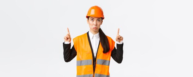 Building construction and industrial concept Disappointed gloomy asian female engineer architect in safety helmet and reflective clothing pouting displeased as looking and pointing fingers up