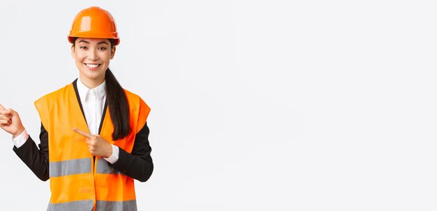 Building construction and industrial concept Smiling asian female architect in safety helmet reflective clothing pointing finger upper left corner showing project on workplace white background