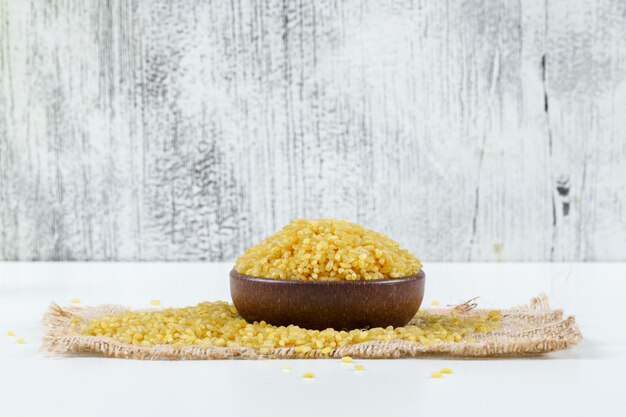 Bulgur cereal in a bowl on piece of sack side view on grunge and white background