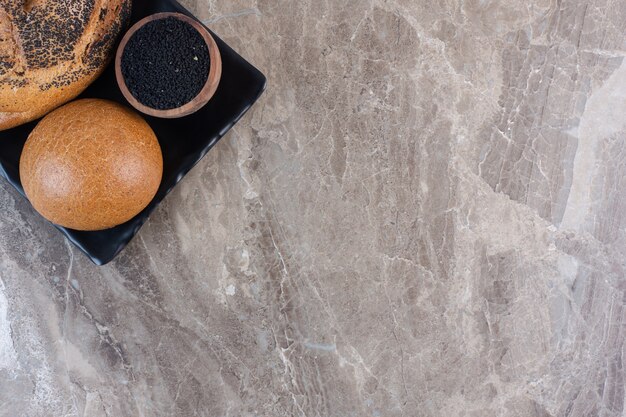 A bun, black sesame coated bread and a small bowl of black sesame seeds on a platter on marble.