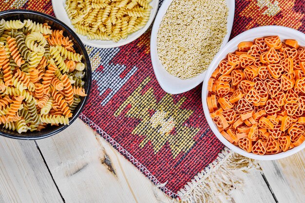 A bunch of colorful uncooked pasta bowls on wooden table with carved cloth.