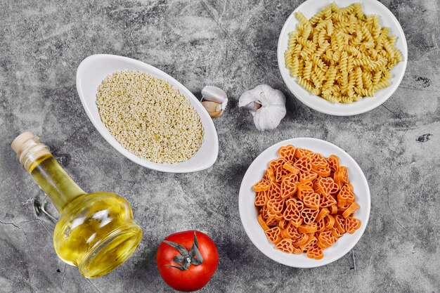 A bunch of different shaped uncooked pasta bowls on marble table with oil, garlic and tomato.