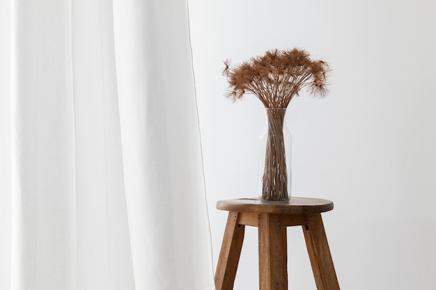 Bunch of dry papyrus plant in a glass vase on a wooden stool by a white curtain