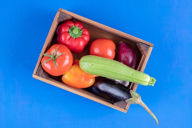 Bunch of fresh ripe vegetables in wooden box on blue surface. 