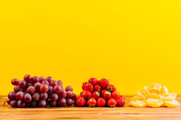 Гроздь винограда; клубника и ломтик апельсина на деревянный стол на желтом фоне