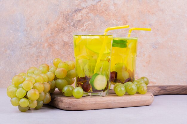 A bunch of green grapes on wooden board with two glasses of juice.