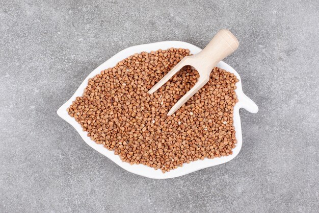 Bunch of raw buckwheat on white plate with wooden spoon