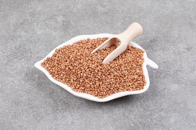 Bunch of raw buckwheat on white plate with wooden spoon