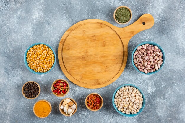 Bunch of spices and bowls of beans, corns around wooden board.