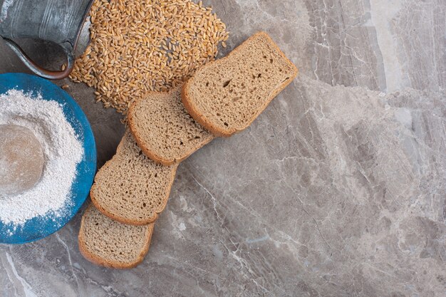 Bundle of bread slices, flour platter and spilled jug of wheat on marble.