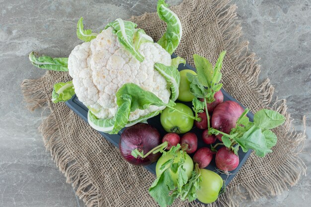 Bundle of red onion, cauliflower, green tomato, turnips and turnip leaves on a black platter on marble.