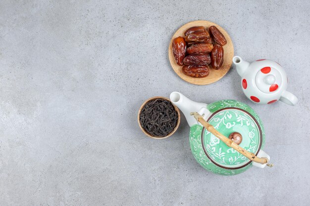 Связка из двух чайников, небольшая миска с чайными листьями и пригоршня фиников на мраморном фоне.