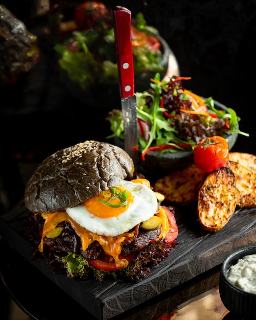 Burger in black bun with fried egg and potatoes.