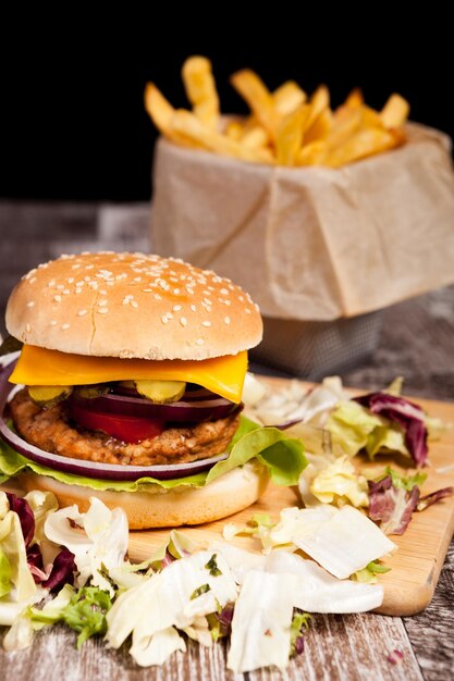 Burger on wooden plate next to fries on black background. Fast food. Unhealthy snack