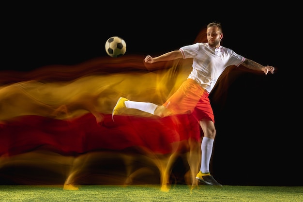 Burning. Young caucasian male football or soccer player in sportwear and boots kicking ball for the goal in mixed light on dark wall. Concept of healthy lifestyle, professional sport, hobby.