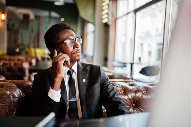 L'uomo afroamericano d'affari indossa un abito nero e occhiali seduto in ufficio e parla al telefono dietro il laptop