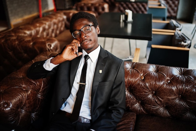 Foto gratuita l'uomo afroamericano d'affari indossa un abito nero e occhiali seduto in ufficio e parla al telefono