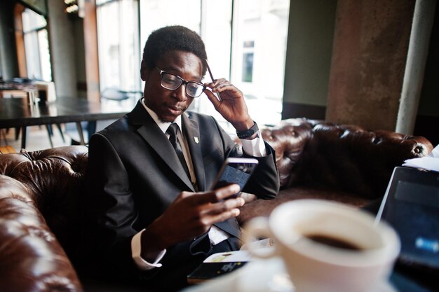 Деловой афроамериканец в черном костюме и очках сидит в офисе с ноутбуком и работает, глядя на мобильный телефон