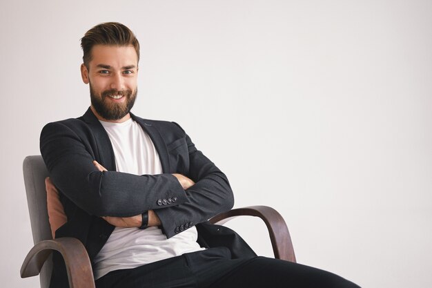 Business, career and success concept. Cheerful stylish young unshaven businessman with happy smile, sitting in armchair against blank wall  with copy space for your information